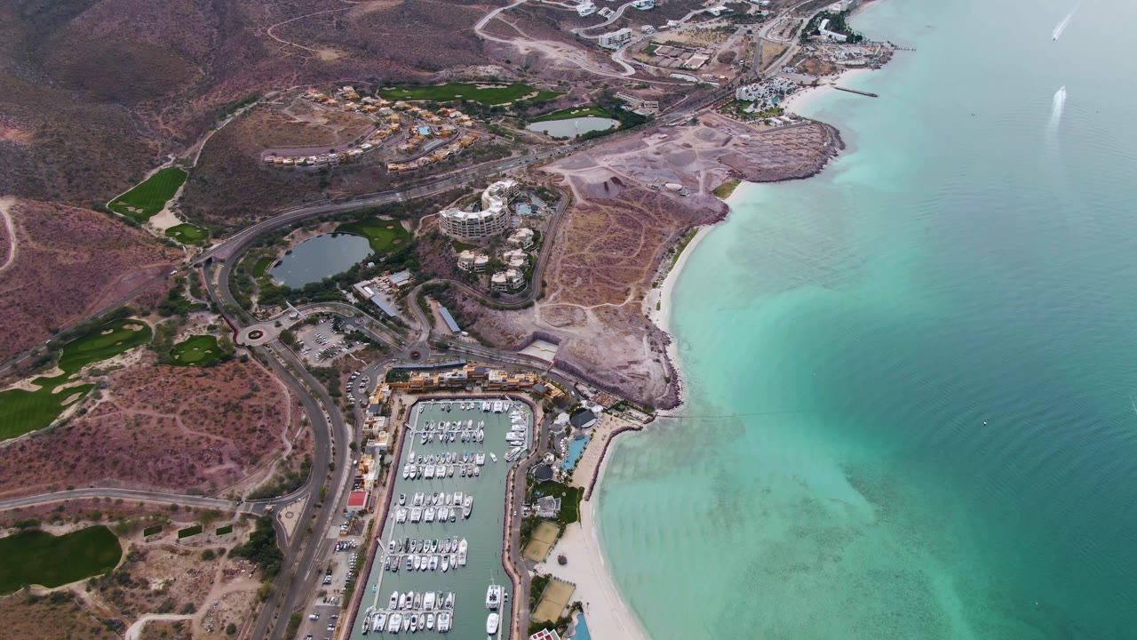 High aerial over a coastal marina, beach, hills, and winding road.
