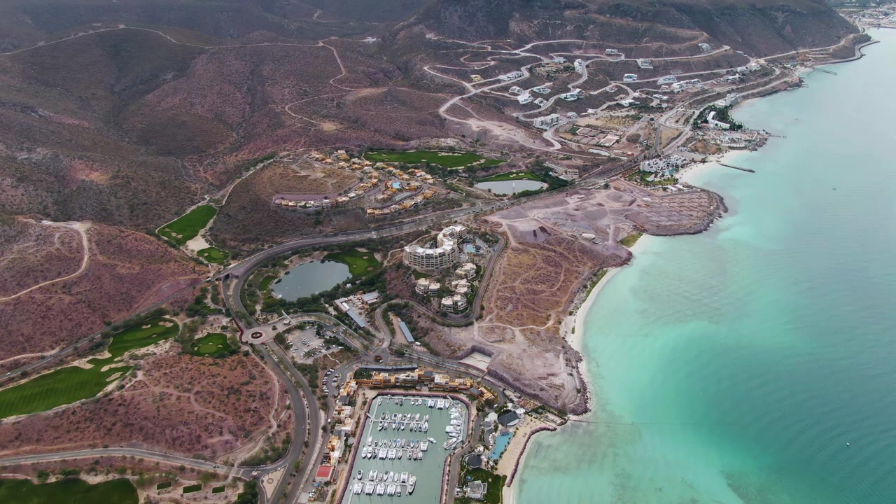 Aerial frame in La Paz, Baja California Sur with the marina lower center, a curved shoreline to the right, dry hillside roads above, green patches inland, and no visible people.