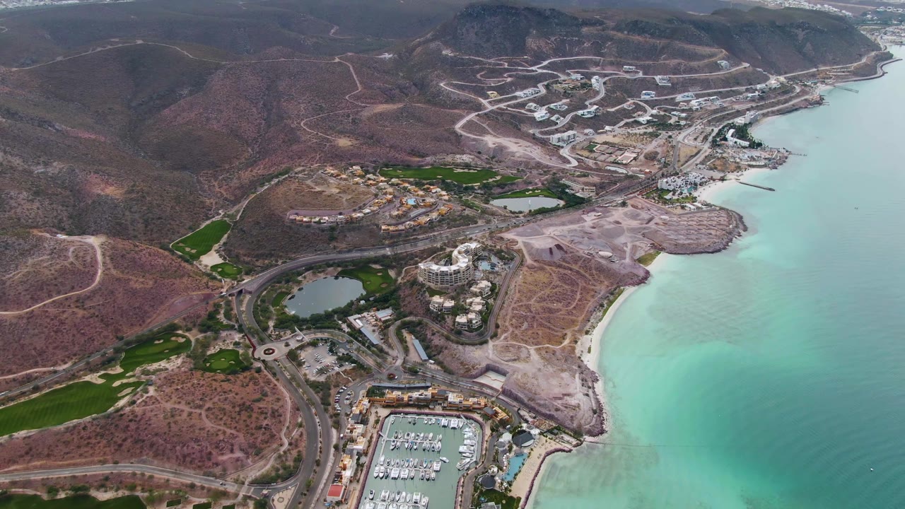 La Paz aerial frame looking across docked boats, roadside buildings, a pale beach edge, turquoise bay water, and dry hills with no visible people.