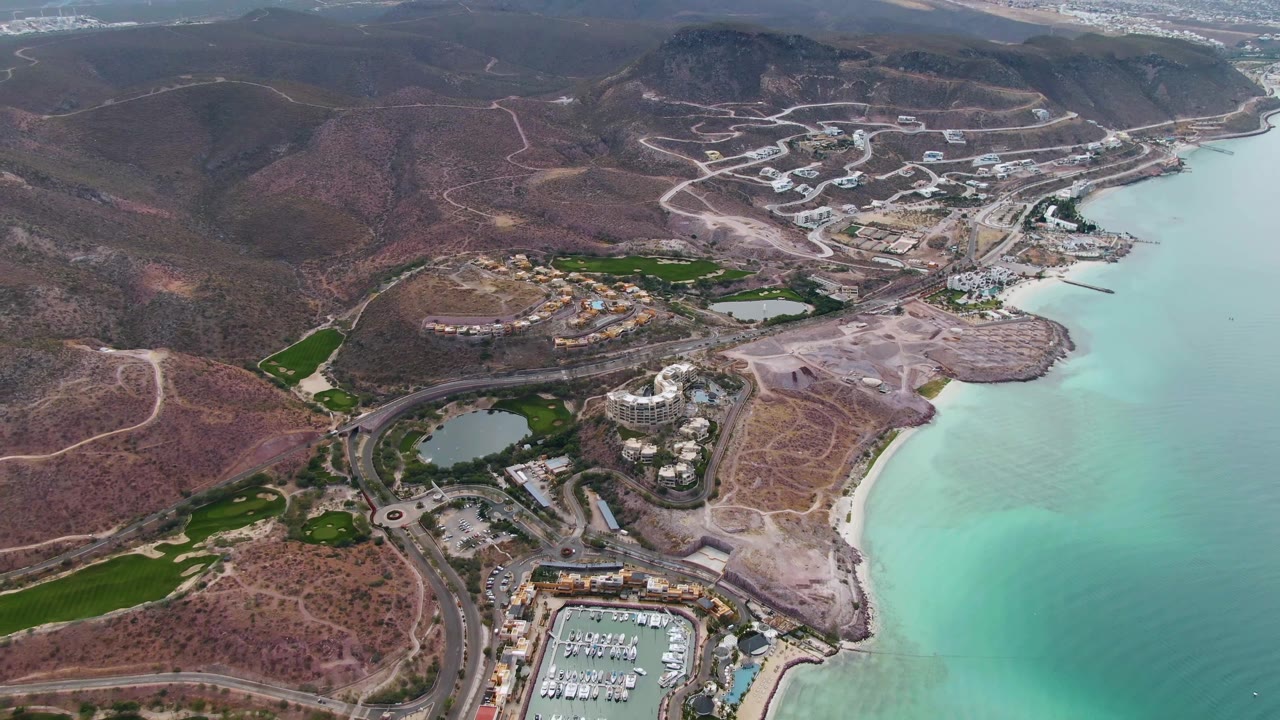 Aerial coastal scene with marina, roundabout roads, green areas, beach, and hills.