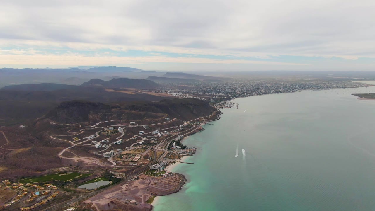 Opening aerial frame over La Paz, Baja California Sur, showing shoreline roads and buildings, green-blue bay water, distant mountains, cloud cover, and no visible people.
