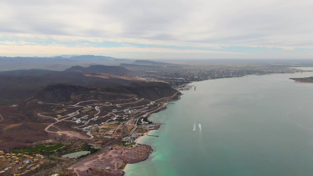 Second aerial frame in La Paz, Baja California Sur, with the boat wake slightly farther across the water, developed shoreline below, mountains in the distance, and no visible people.