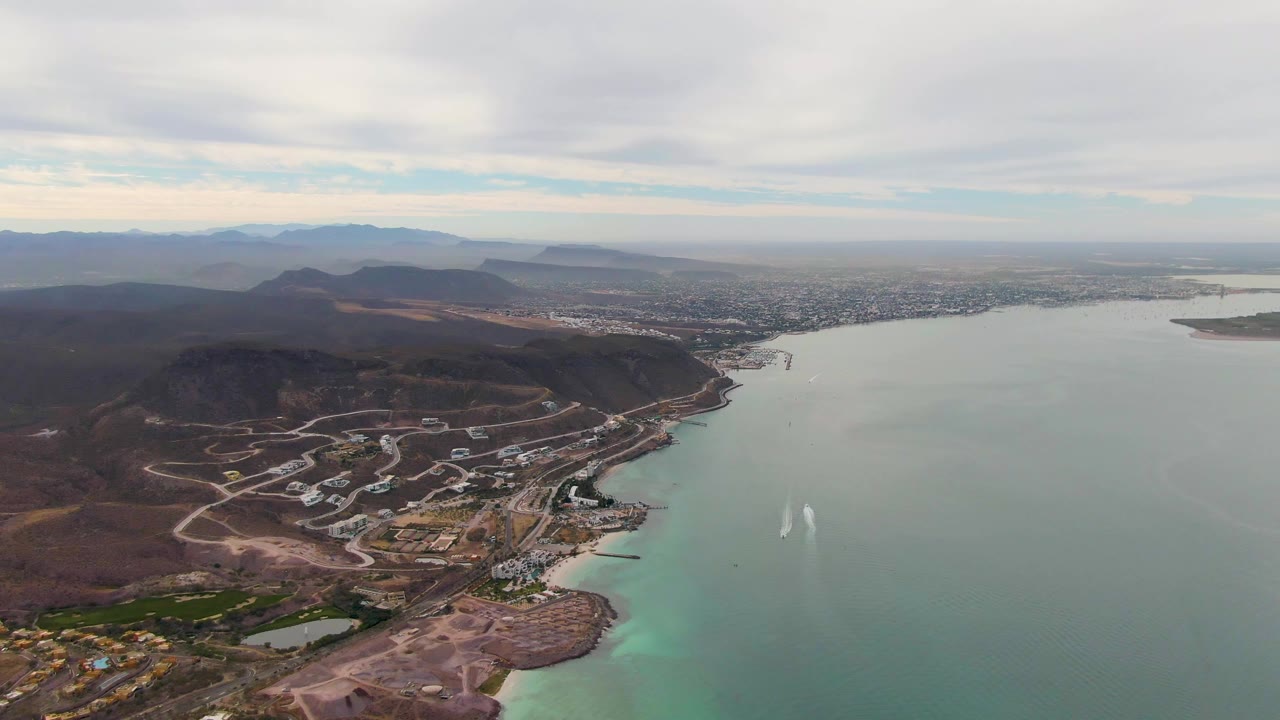 Third frame over La Paz, Baja California Sur, showing broad open water, a faint twin wake, shoreline construction roads, low buildings, mountain ridges, and no visible people.