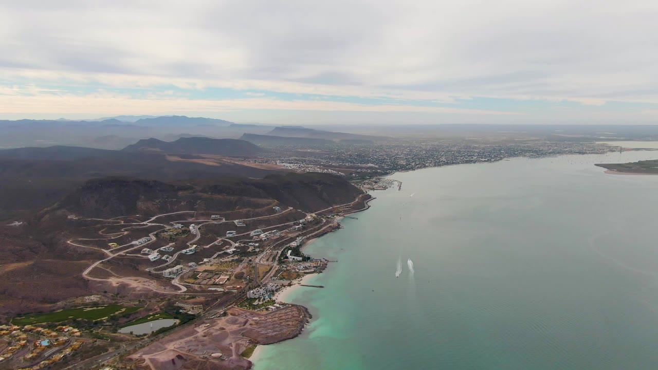 Aerial view of brown hills, coastal roads, buildings, green water, and a fading boat wake.