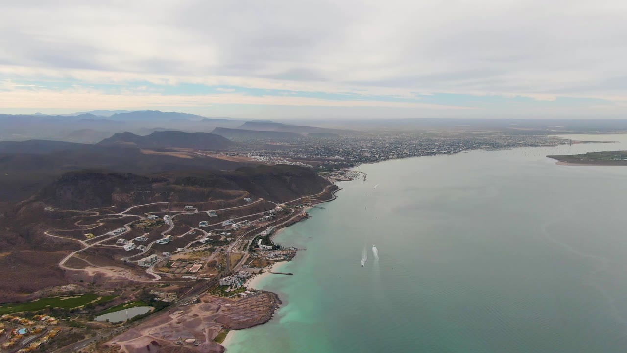 Opening aerial frame over La Paz, Baja California Sur, with dry hills and shoreline roads in the foreground, muted bay water to the right, distant city on the horizon, and no visible people.