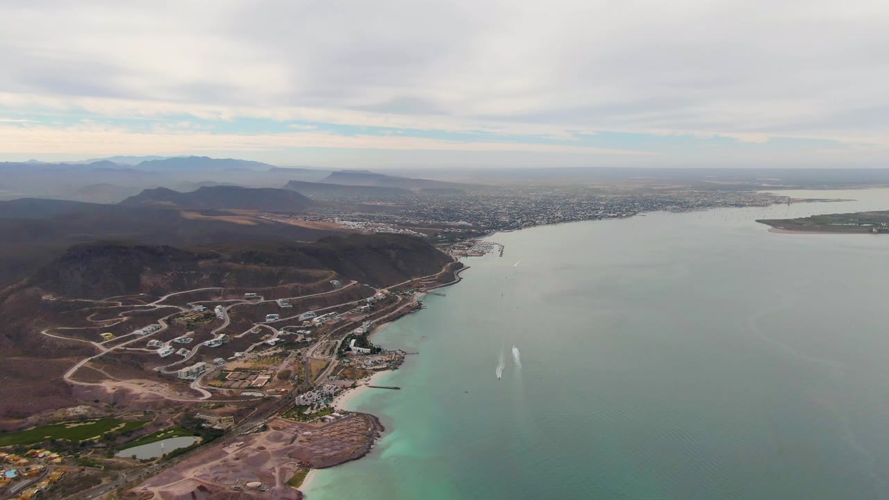 Mid-clip aerial frame in La Paz showing dark brown hills and road patterns at left, broad green-blue bay water at right, city and mountains under an overcast sky, and no visible people.