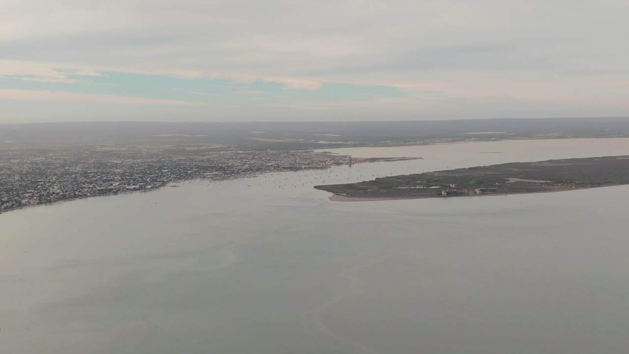 Wide aerial frame over La Paz, Baja California Sur, with the city spread along the left shoreline, calm water in front, a low landmass to the right, and no visible people.