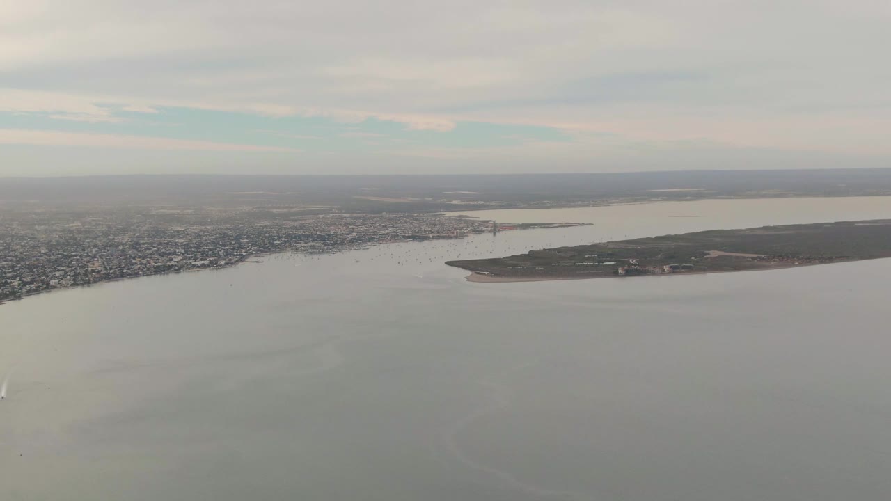 Aerial view over La Paz water with the city along the far shore, a low island or peninsula to the right, a small white sailboat wake near the lower center, and no visible people.