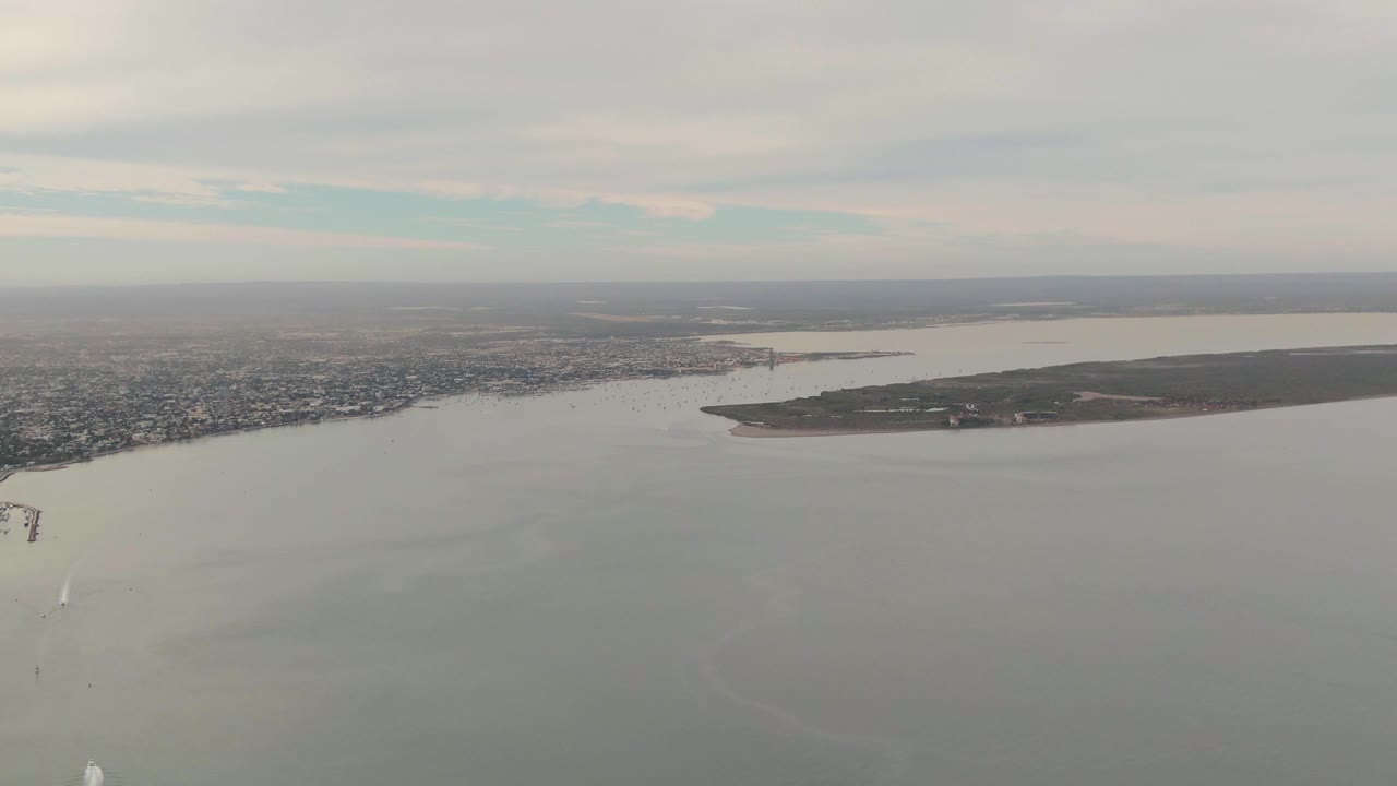 Wide aerial frame in La Paz with gray-blue water in the foreground, distant urban shoreline across the horizon, the right-side landmass more centered, several pale boat wakes, and no visible people.