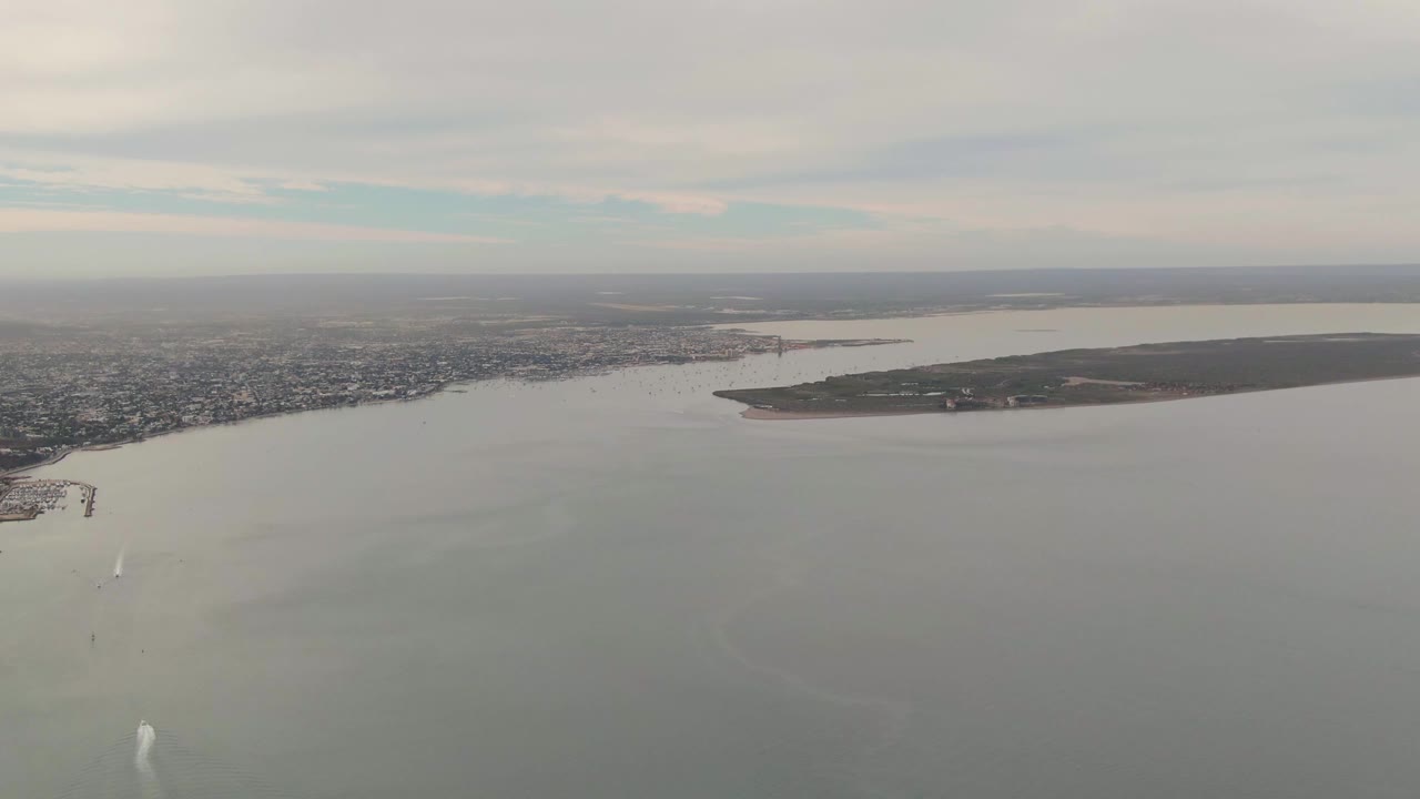 Aerial view of La Paz shoreline, water, and low island with a visible sailboat wake.