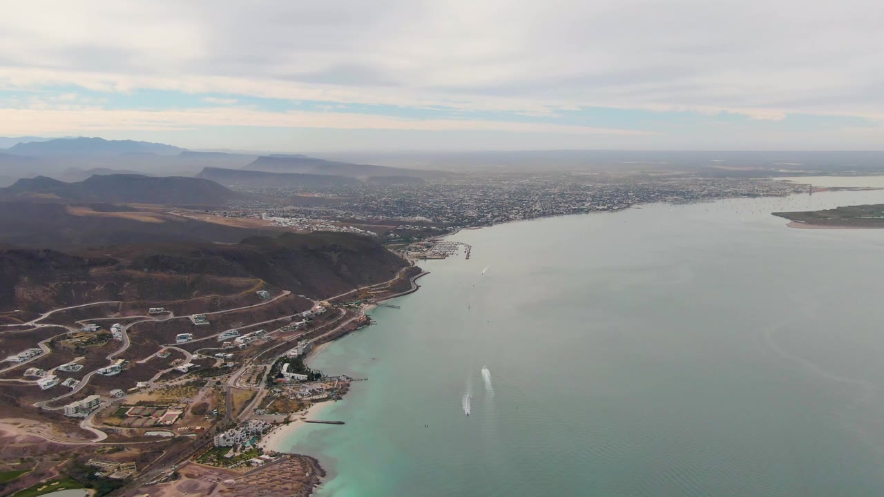 Aerial frame over La Paz, Baja California Sur showing hillside roads and buildings at left, pale water across the center, distant city and mountains on the horizon, and no visible people.