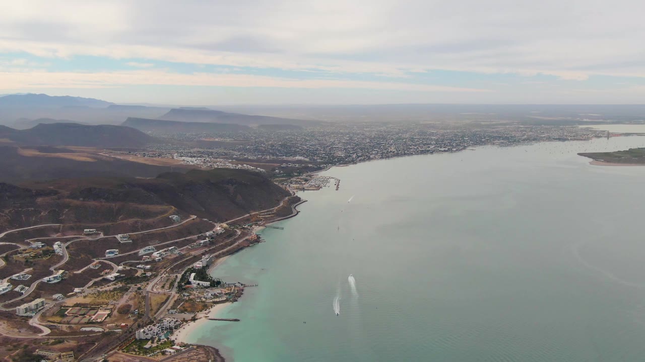 Aerial view in La Paz with winding hillside roads and waterfront structures in the lower frame, broad green-blue water to the right, city across the bay, distant mountains, and no visible people.