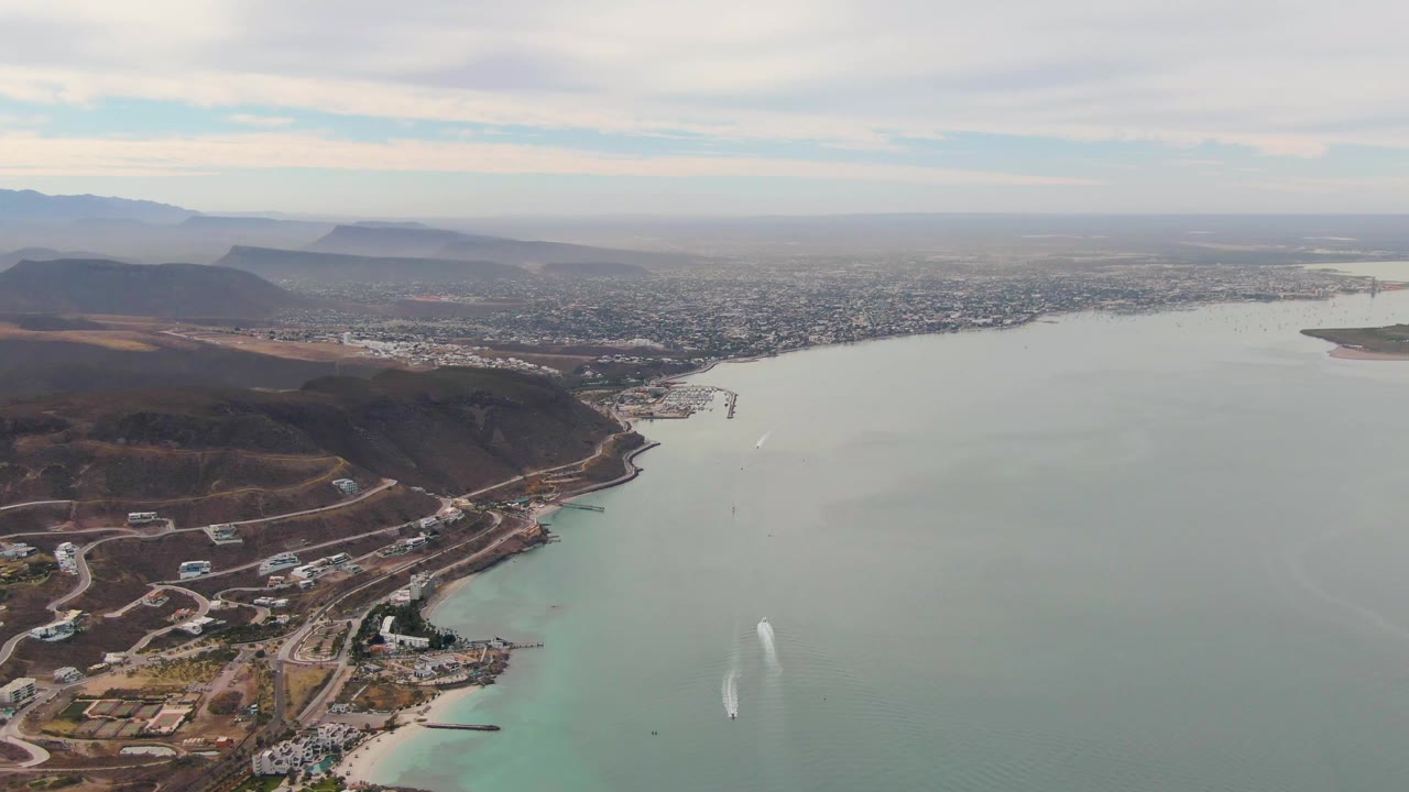 La Paz aerial frame showing the hillside and roads shifted farther left, open water filling the foreground and right side, the city stretching across the bay, mountains behind it, and no visible people.