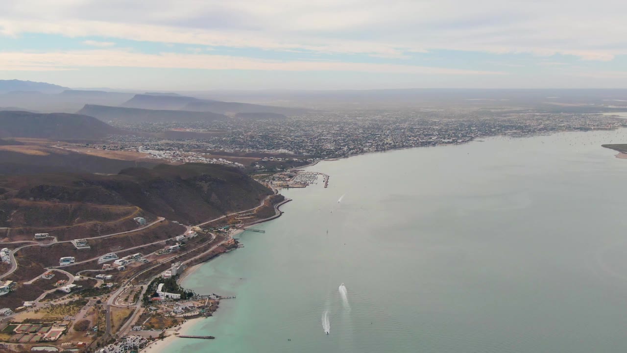 Aerial frame with hillside development at left, bay water at right, and distant urban area under clouds.