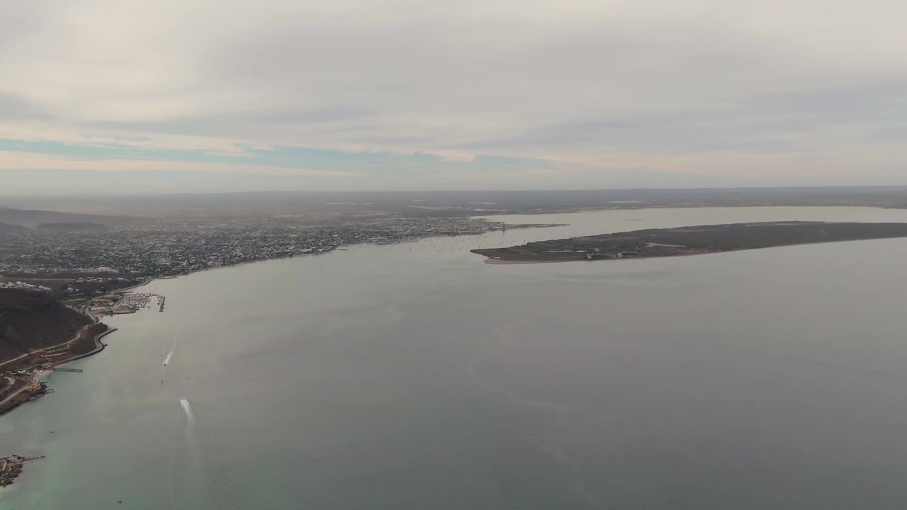 Aerial view of coastal water, boat wakes, city shoreline, and island under clouds.