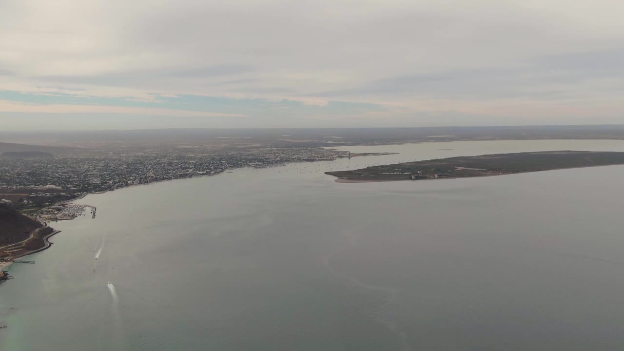 Aerial view over La Paz with pale coastal water in the foreground, a narrow white boat trail near the lower center, city shoreline beyond, island landmass to the right, and no visible people.