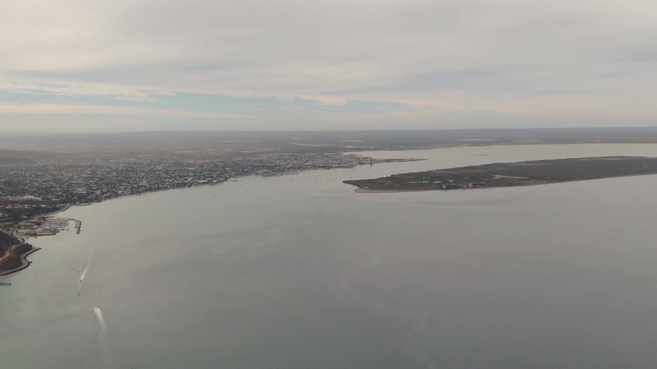 Mid-clip aerial frame of La Paz coastal waters with a white wake lower in frame, the urban shoreline stretching across the background, island terrain on the right, soft overcast light, and no visible people.