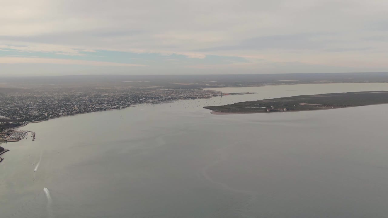 Aerial frame over La Paz where the shoreline sits higher in the composition, a white wake enters from the lower left, the island spans the right side, the sky remains cloudy, and no visible people appear.