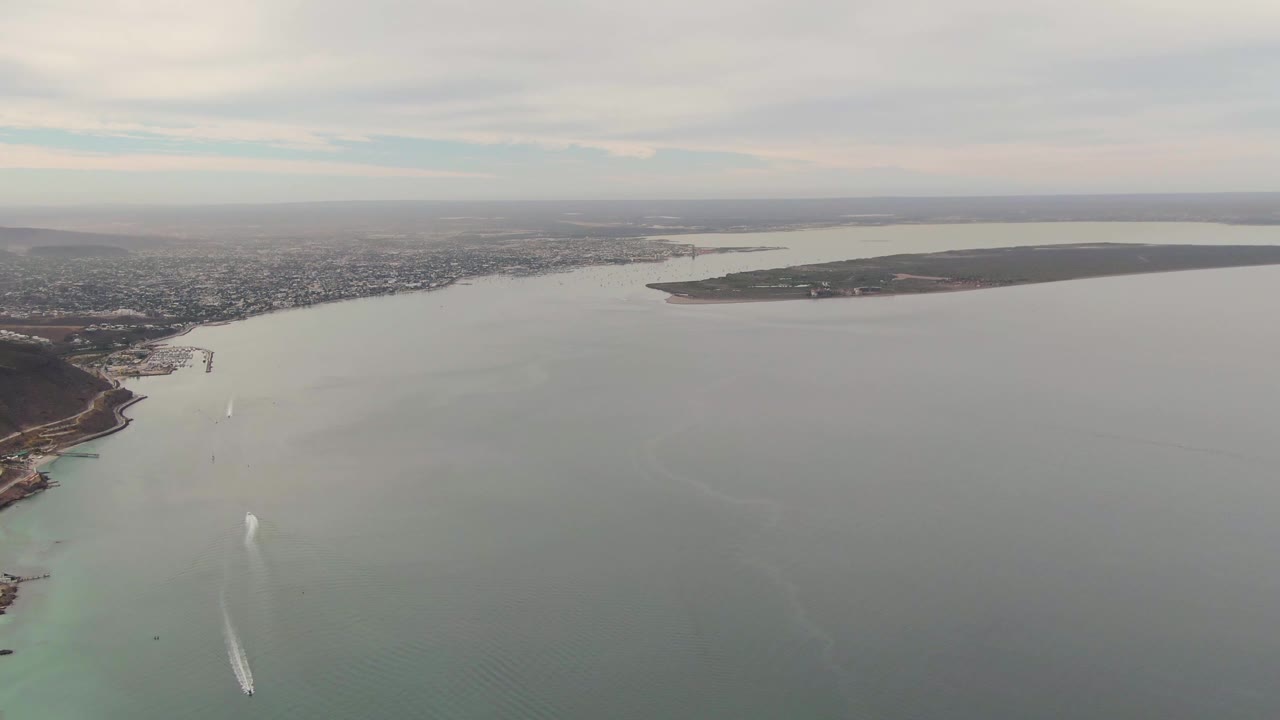 Wide aerial frame over La Paz, Baja California Sur, Mexico, with gray-green bay water, turquoise shallows near the coast, a small white boat wake, distant urban shoreline, and no visible people.