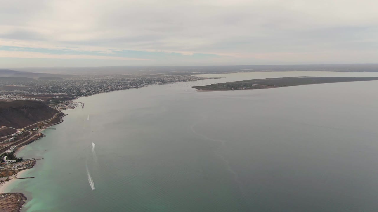 Wide drone-like frame of La Paz bay with the wake centered lower in the water, a brown coastal point at right, pale turquoise shallows along the edge, distant developed land, and no visible people.