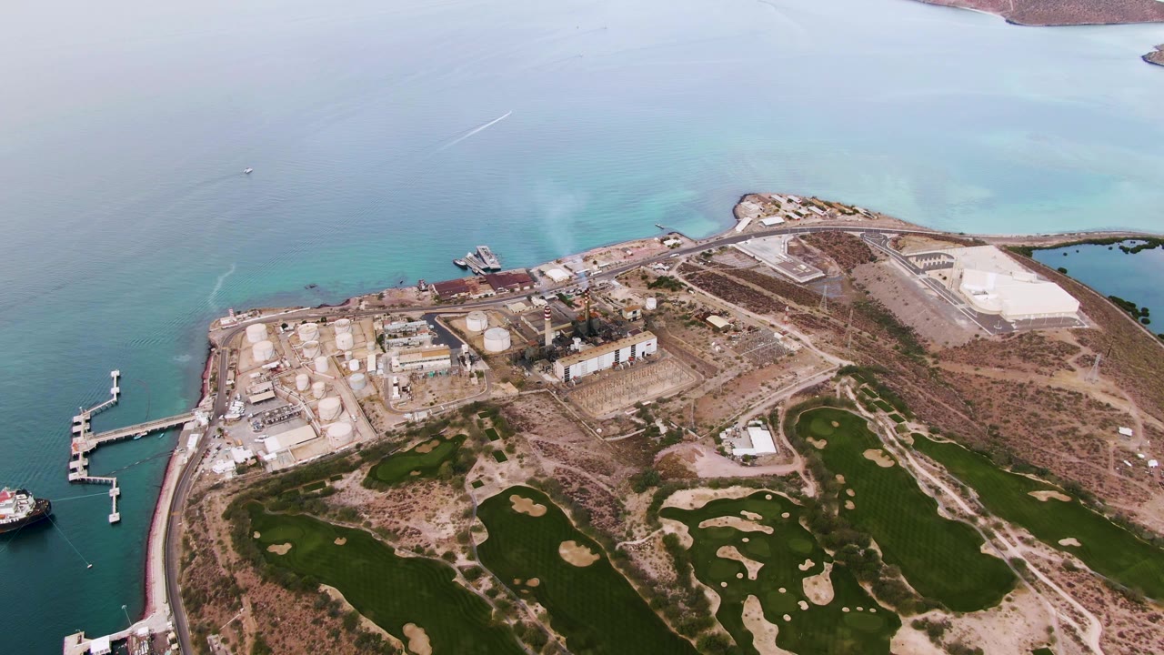High aerial frame over La Paz, Baja California Sur showing industrial tanks and docks along turquoise water, golf fairways and sand traps below, and no visible people.