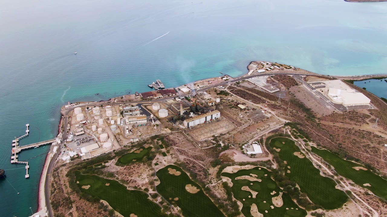 Aerial frame in La Paz, Baja California Sur with the industrial site centered, pale turquoise water behind it, green golf course areas in the lower frame, and no visible people.