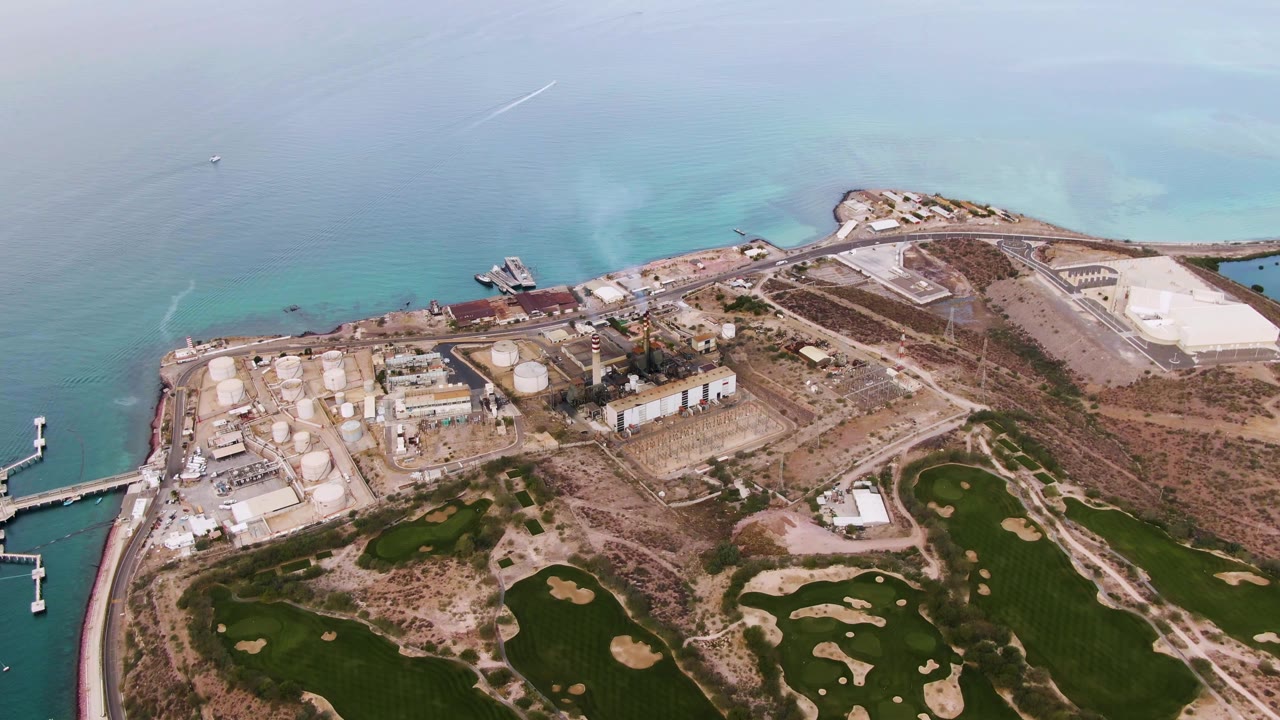 High aerial view of La Paz, Baja California Sur showing coastal industrial buildings and tanks, bright shallow water, golf course greens, sand bunkers, and no visible people.