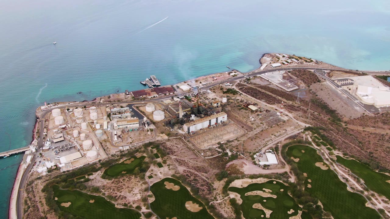 Aerial view of industrial tanks and dock beside calm water with golf course in foreground.