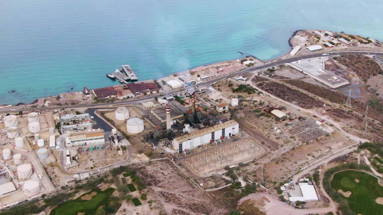 Aerial frame over La Paz, Baja California Sur, showing storage tanks, a chimney with smoke, a coastal road, sandy lots, turquoise water, and no visible people.