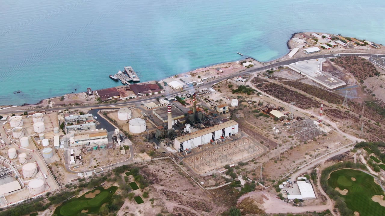Aerial frame over La Paz, Baja California Sur, showing the coastal road crossing behind the plant, smoke above the chimney area, water color bands offshore, and no visible people.