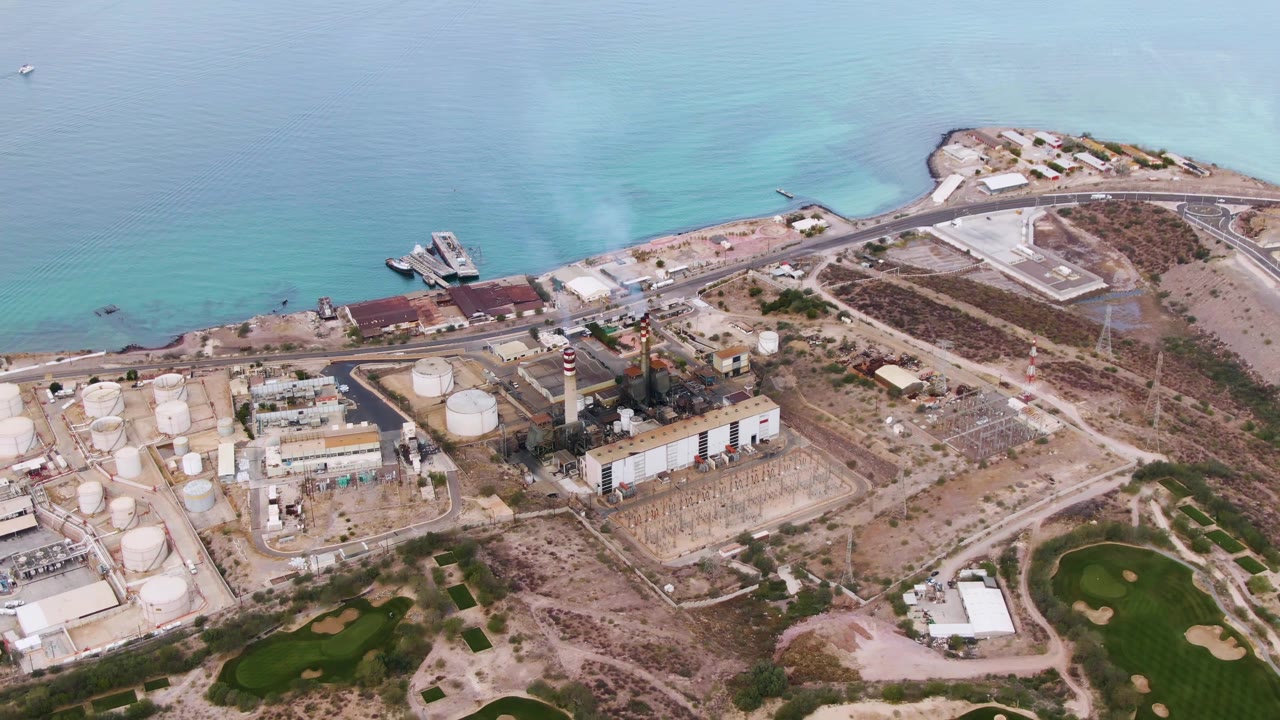 Aerial shot of industrial tanks, smoke, beach road, and green grass areas.