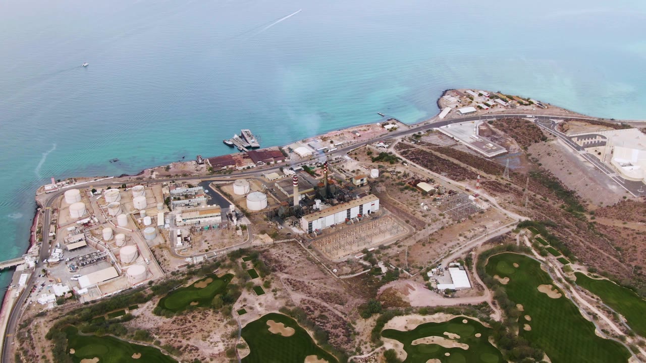 Aerial view over La Paz, Baja California Sur, showing a coastal industrial site, green fields, pale turquoise water, and no visible people at the start of the clip.