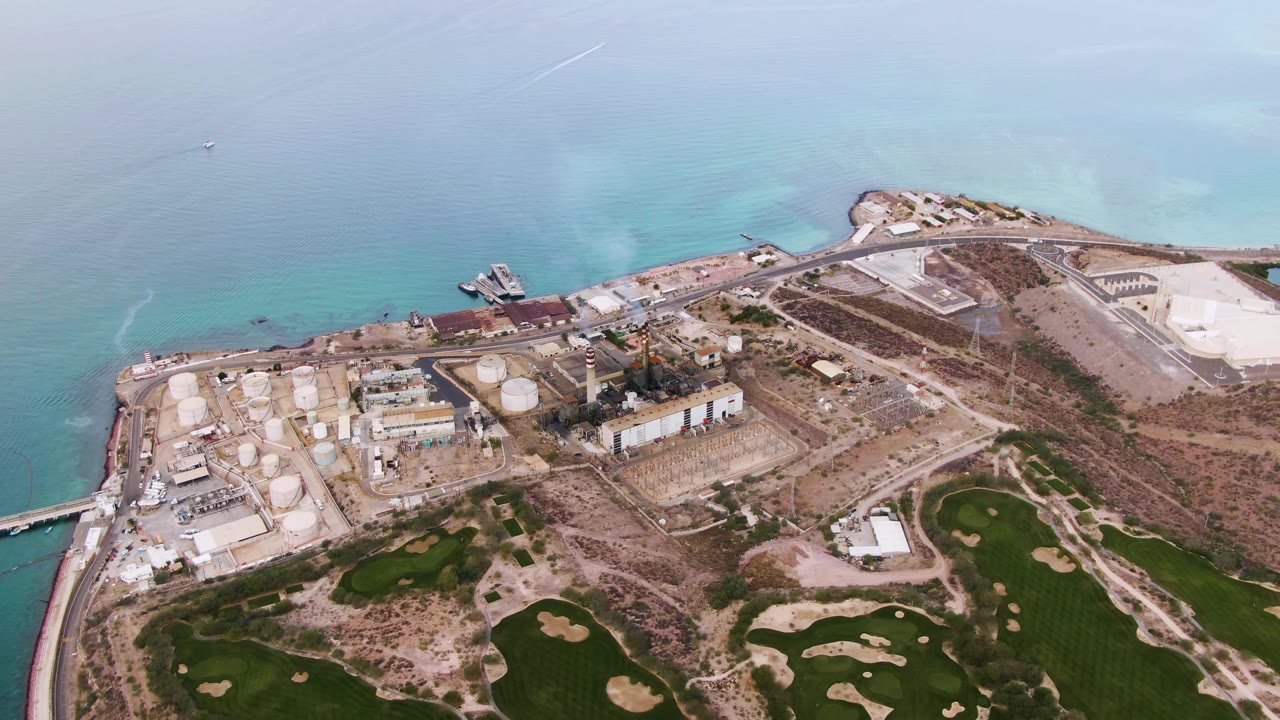 A wide aerial frame in La Paz, Baja California Sur, shows storage tanks near the shore, open water above, green fields below, and no visible people.