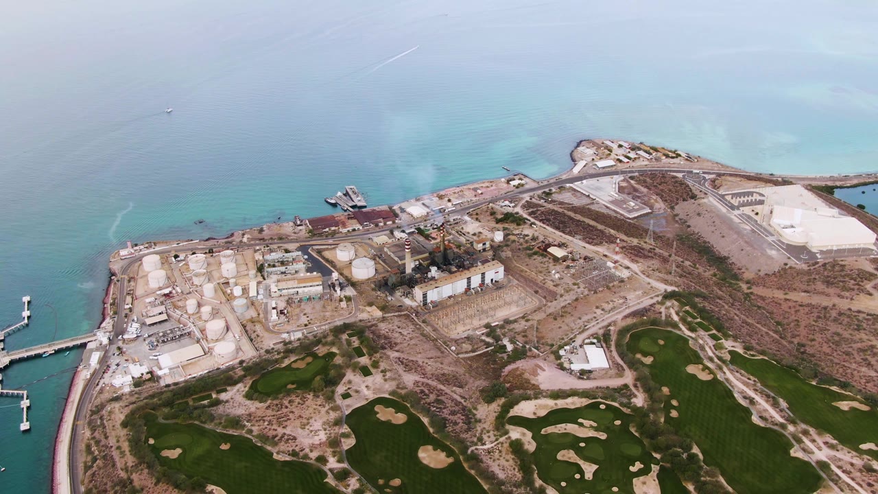 Aerial view in La Paz, Baja California Sur, with dock arms more visible at left, industrial tanks near shore, green fields at bottom, and no visible people.