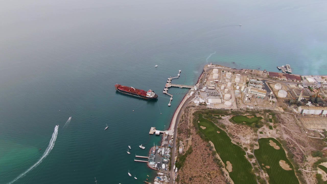 Aerial frame over La Paz, Baja California Sur, showing a red cargo ship near docks, teal water, shoreline tanks, green golf fairways, and no visible people at the start of the clip.