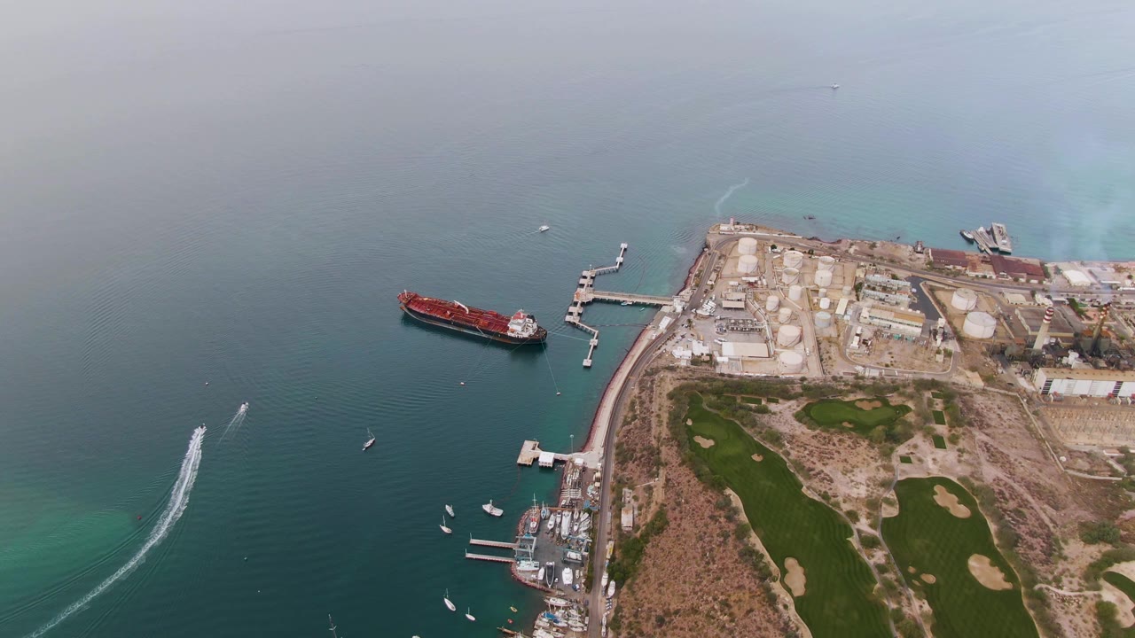 Aerial frame over La Paz with the red cargo ship centered left of the docks, a curved white boat wake on the lower left, industrial storage tanks onshore, and no visible people.