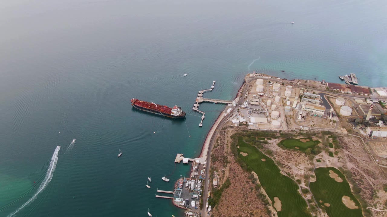 Aerial frame in La Paz showing the red cargo ship slightly left of center, small boats scattered below it, pale industrial ground and tanks on the right, golf course greens at lower right, and no visible people.