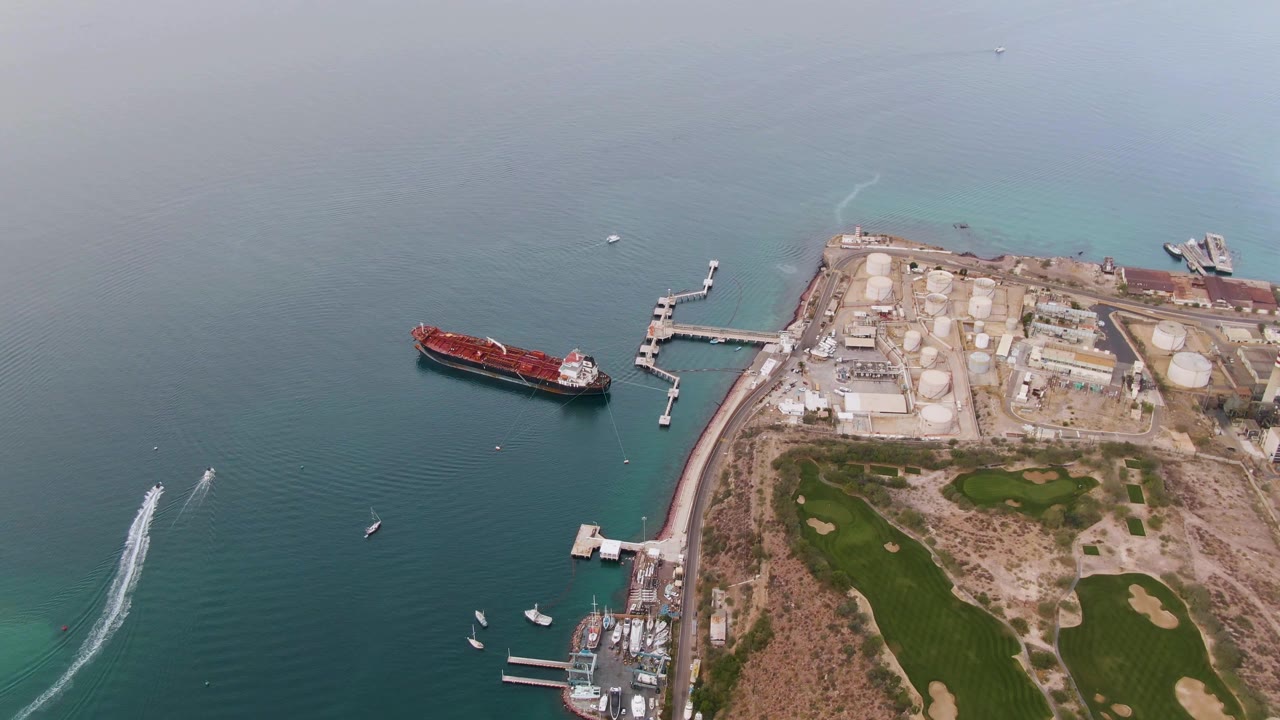 Aerial coastal view with red cargo ship, boat wake, docks, tanks, and golf course.
