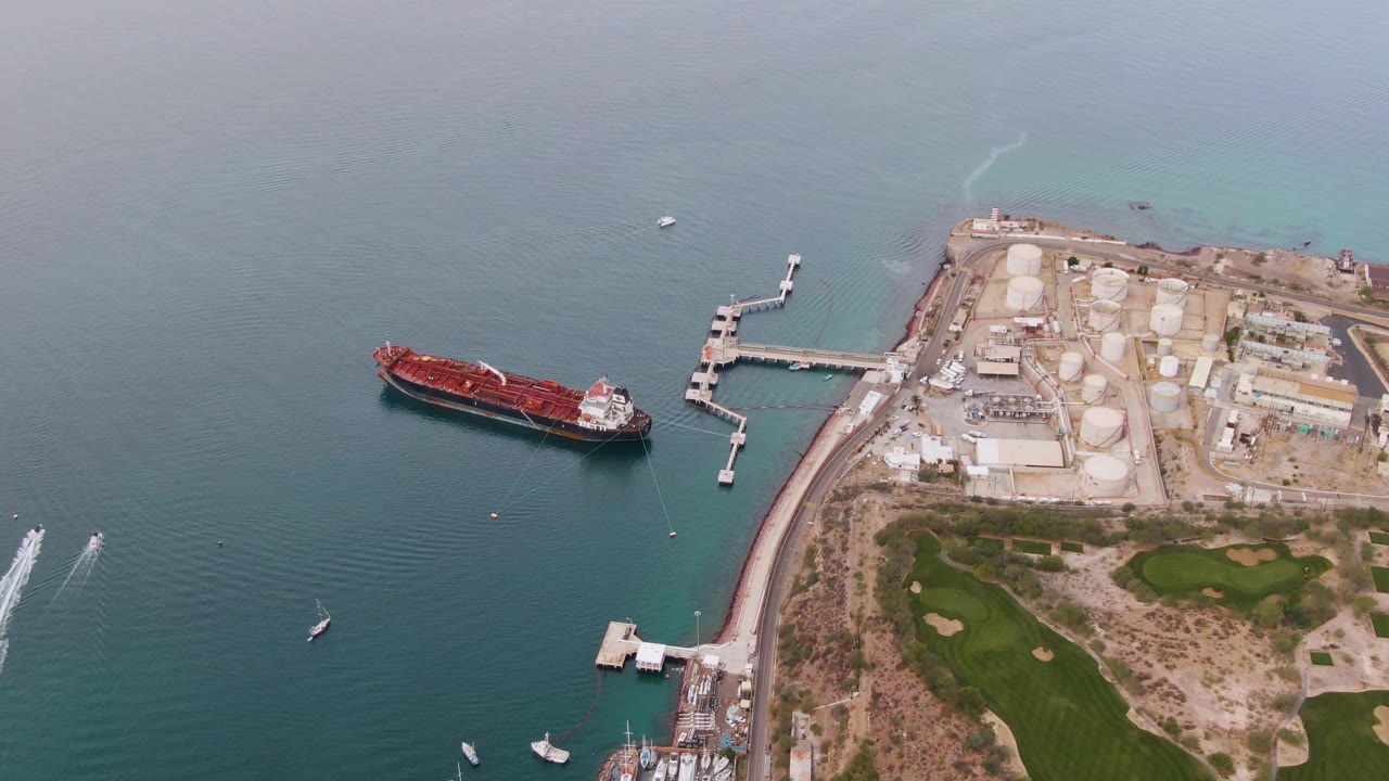 Aerial view of a red cargo ship beside a coastal industrial dock and storage tanks.