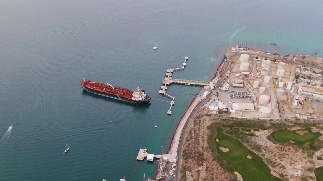 Aerial view over La Paz, Baja California Sur with the red cargo ship left of the dock, industrial tanks to the right, a small boat lower left, and no visible people.