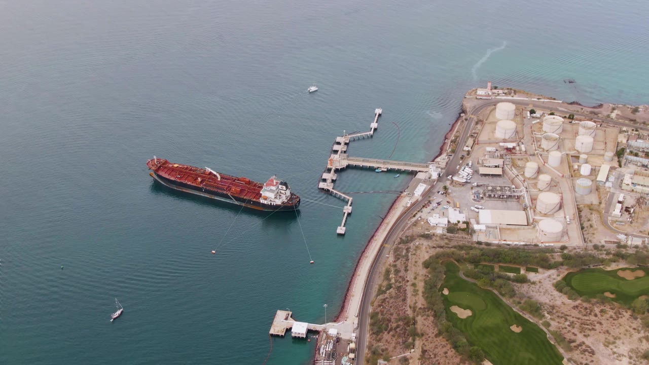 Aerial frame in La Paz, Baja California Sur showing the cargo ship aligned with the dock, white tank structures on shore, faint wakes in the upper water, and no visible people.