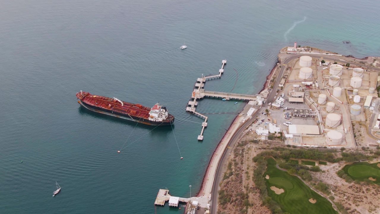 Aerial view from La Paz, Baja California Sur with the red cargo ship slightly lower in frame, the dock and storage tanks to the right, green land at bottom right, and no visible people.