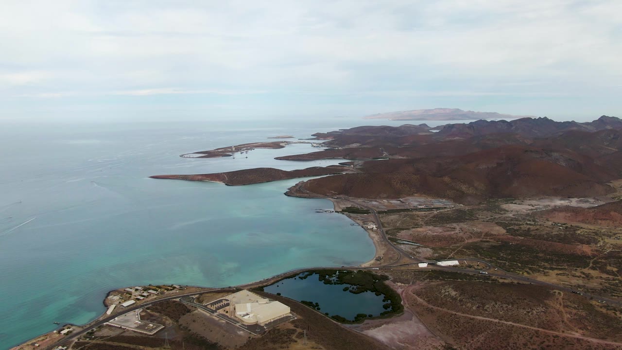 Aerial frame over La Paz, Baja California Sur showing turquoise coastal water beside dry mountain ridges, a road, a dark pond, distant islands, cloudy sky, and no visible people.