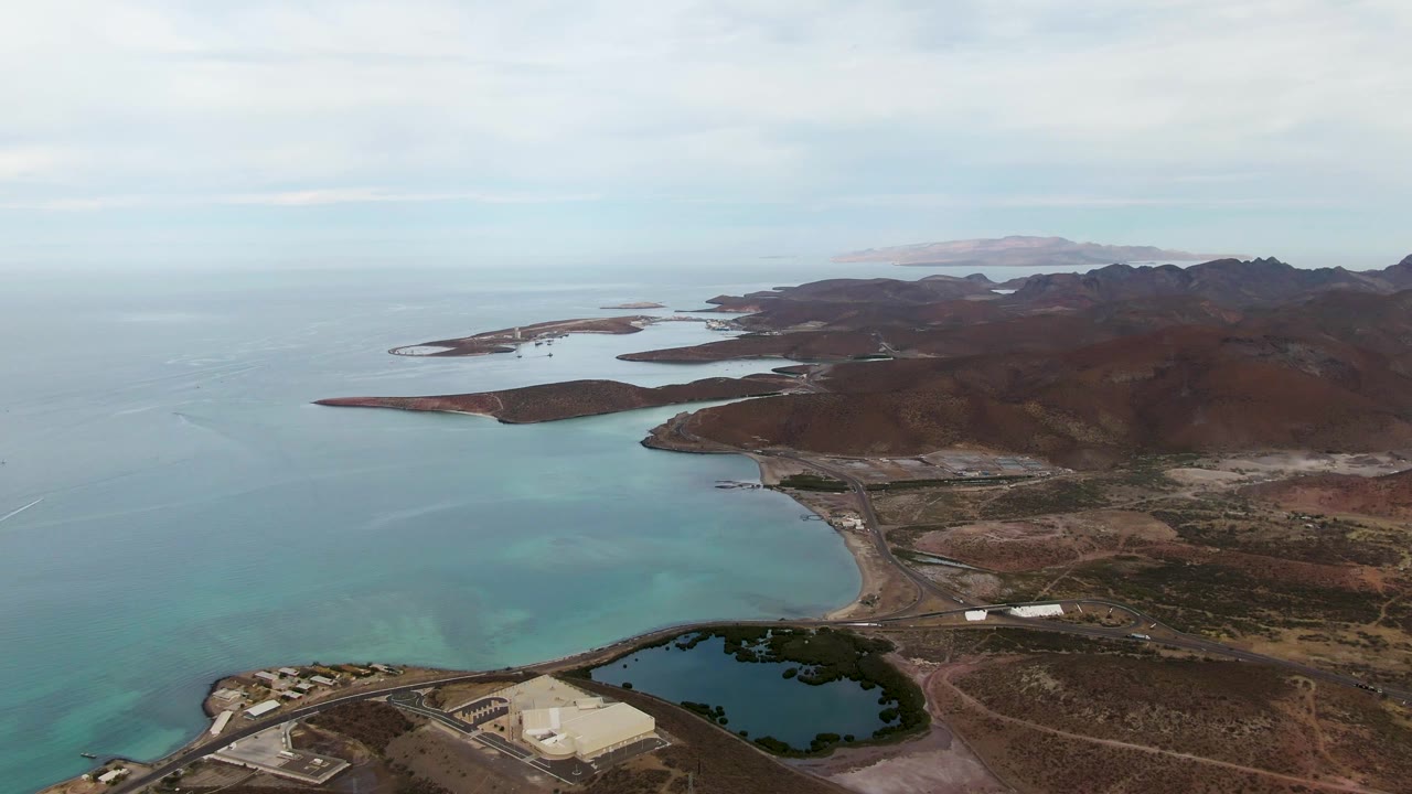 Drone frame over La Paz with the shoreline road and dark pond near the lower frame, pale turquoise water on the left, brown ridges on the right, distant islands, and no visible people.