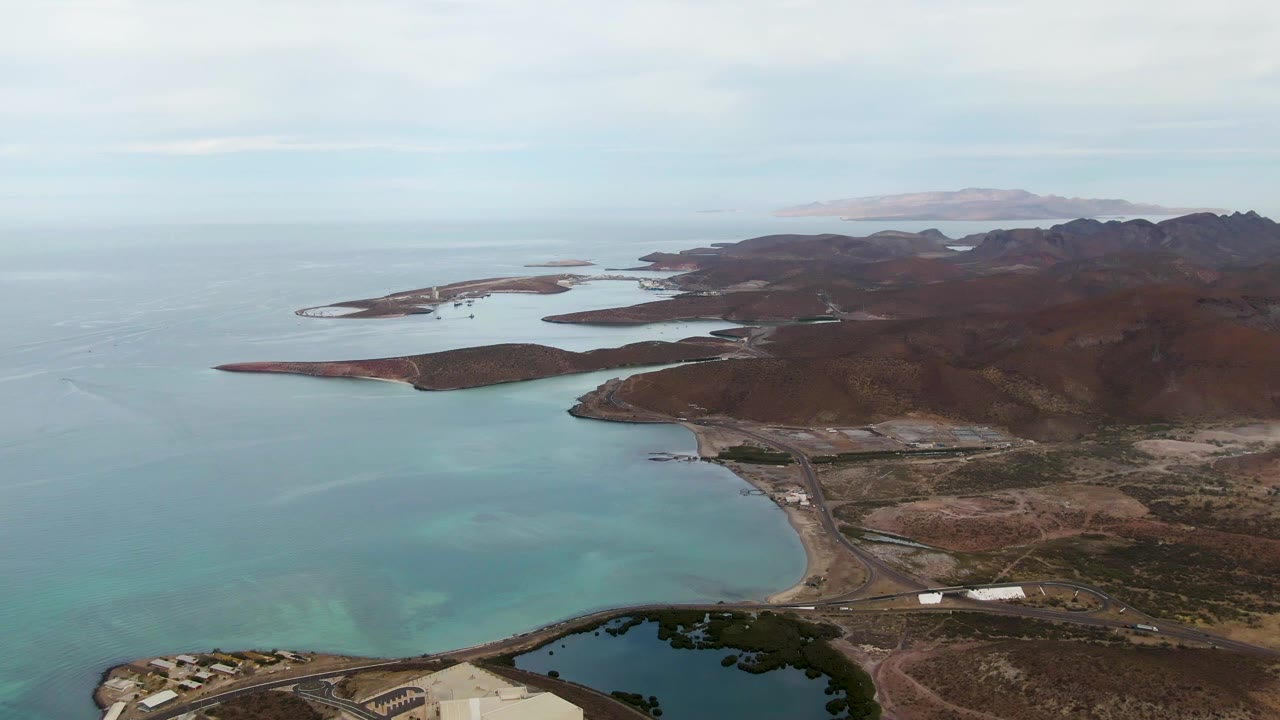Aerial coastline view with broad bay, rocky ridges, road corridor, and distant islands.