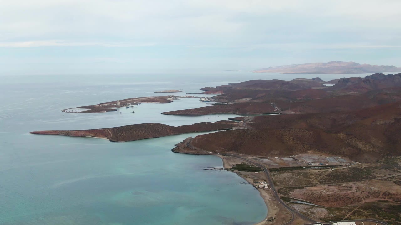 Aerial frame over La Paz, Baja California Sur showing rocky islands, pale turquoise shallows, dry coastal land, a small shore structure area near the lower right, and no visible people.