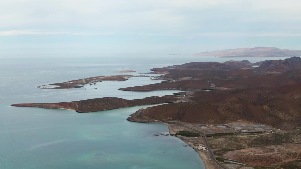 Aerial frame over La Paz, Baja California Sur with calm blue water in the foreground, low rocky islands across the midground, brown hills to the right, hazy distant land, and no visible people.