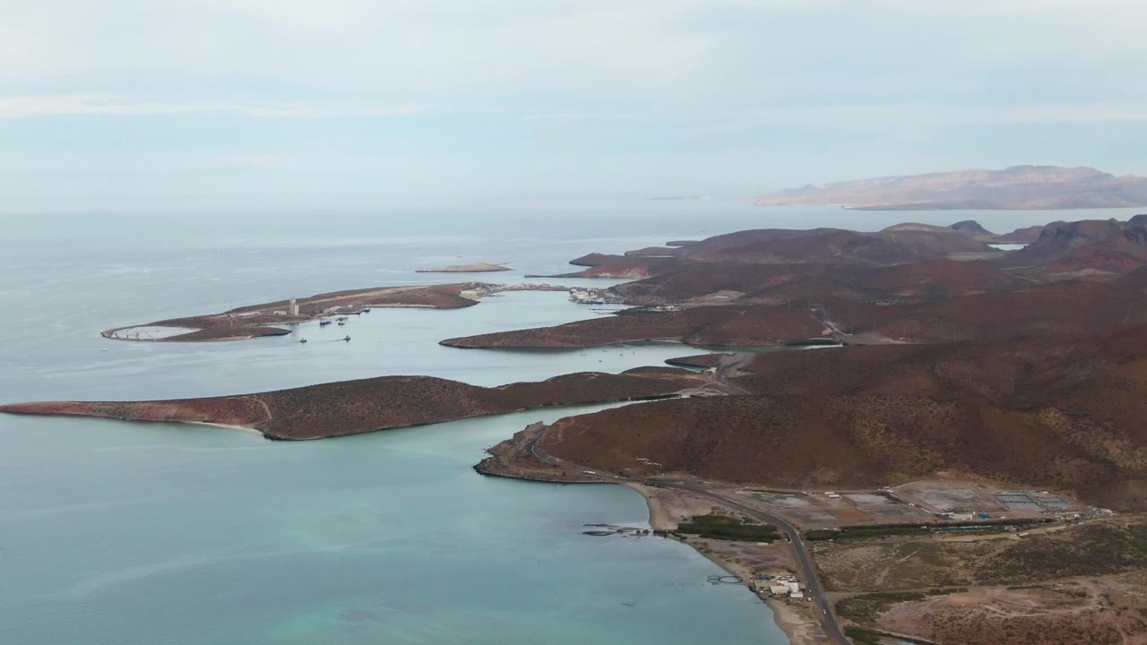 Aerial frame over La Paz, Baja California Sur showing a long low island beside turquoise shallows, darker rocky hills on the right, faint land on the horizon, and no visible people.