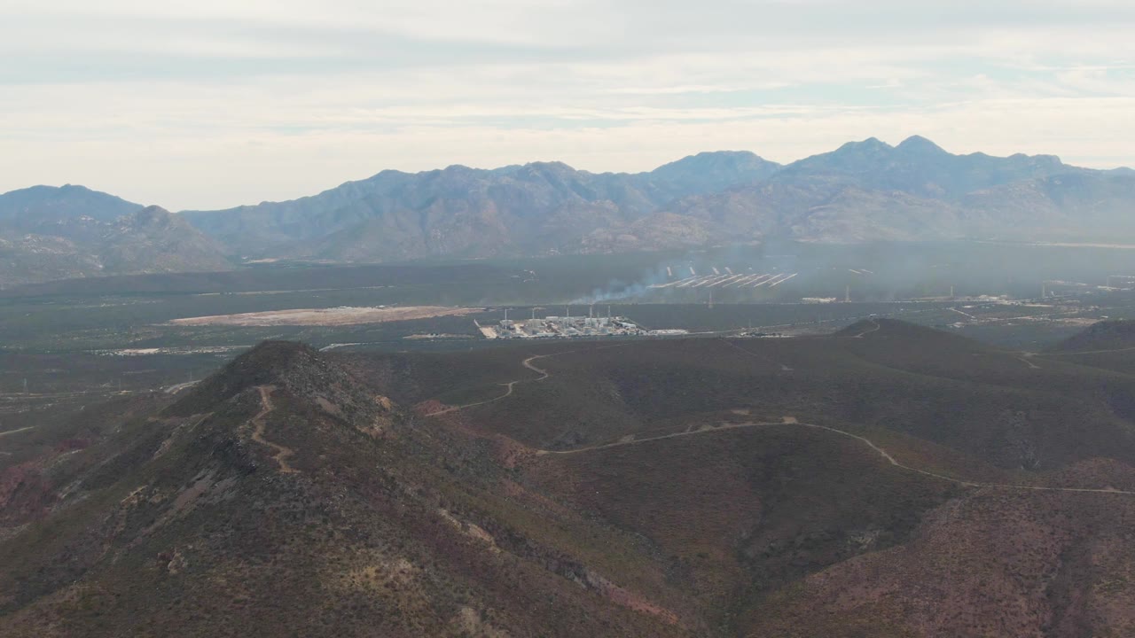 At one second, the La Paz valley view holds steady with the foreground ridge lower left, a pale smoke plume near the settlement, hazy mountains beyond, and no visible people.