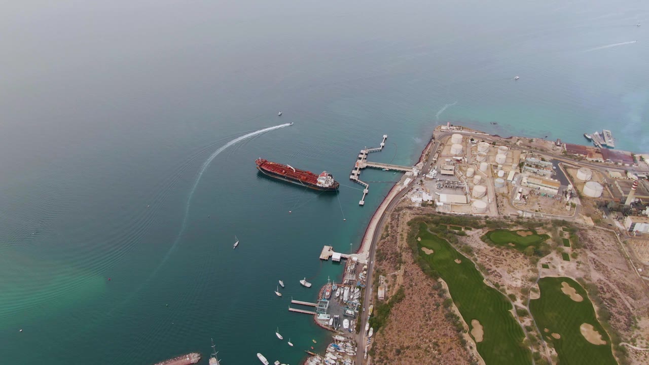 High aerial frame over La Paz, Baja California Sur, Mexico, showing a red cargo ship on blue-green water, a curved speedboat wake, industrial tanks, marina docks, golf course greens, and no visible people.